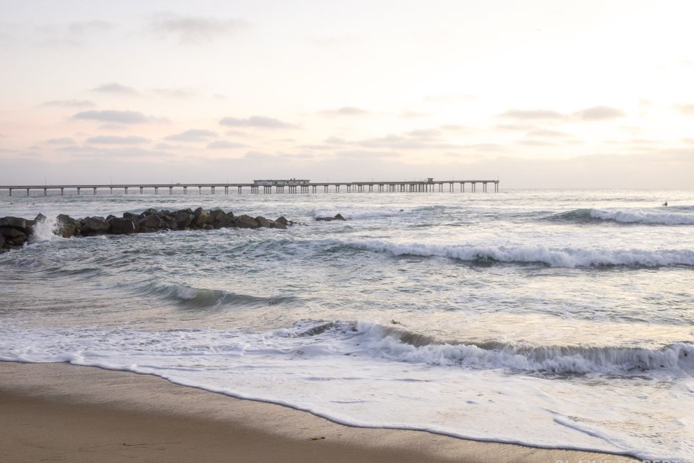 Gentle waves wash onto the sand at sunset, with a rocky jetty cutting into the surf and Ocean Beach Pier stretching across the horizon beneath a soft, cloudy sky.