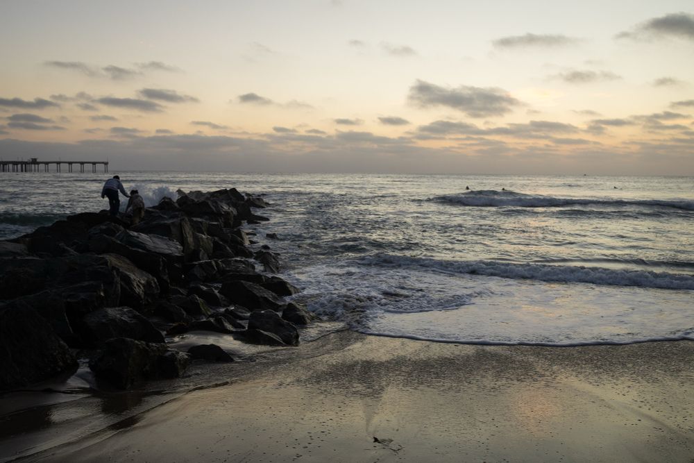 Two people climb carefully across large jetty rocks at the edge of the ocean during sunset. Gentle waves roll in under a sky filled with soft clouds, and the wet sand reflects the pastel light. A distant pier stretches into the sea on the left, while a few surfers sit quietly in the water.