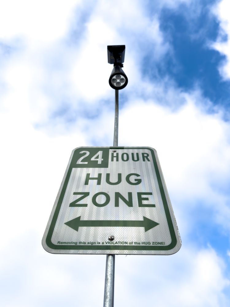 A street-style sign on a pole reads “24 HOUR HUG ZONE” with arrows pointing left and right. The sign is centered against a bright, partly cloudy sky. A small streetlamp is mounted above the sign.