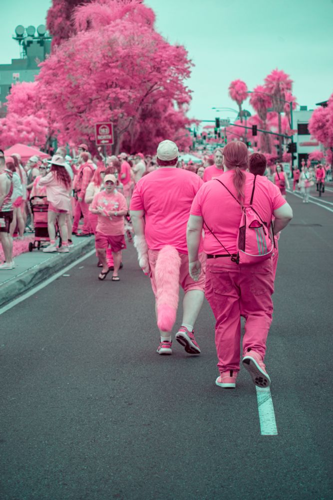 Two people walk side by side down a city street during a Pride parade, both dressed in bright pink clothing. One wears a pink fuzzy tail, and the other carries a pink drawstring backpack. The surrounding crowd and trees appear vibrant pink due to infrared photography, creating a surreal, celebratory atmosphere. The white road line leads into a dense background of palm trees and festivalgoers.