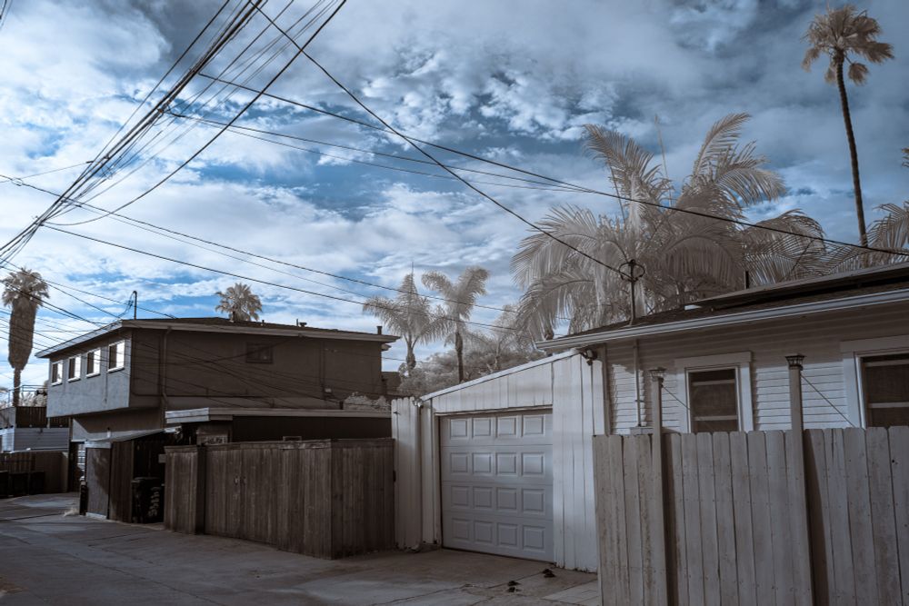 Infrared image of an alleyway lined with fences, a detached garage, and utility poles. Tall palm trees appear pale and frosted against a textured sky with streaks of clouds and power lines crossing overhead.