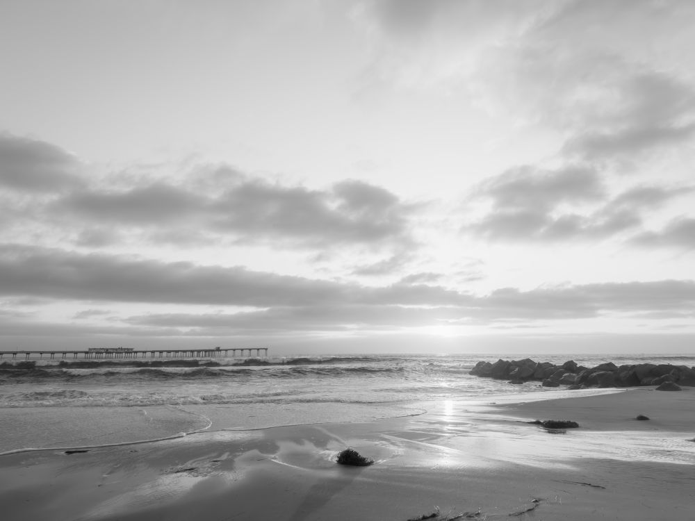 Black and white photo of a calm beach at low tide with gentle waves and wet sand reflecting light. A rocky jetty extends from the right, and a long pier stretches out into the ocean on the left. The sky is filled with scattered clouds, and the sun is low on the horizon, creating a serene atmosphere.