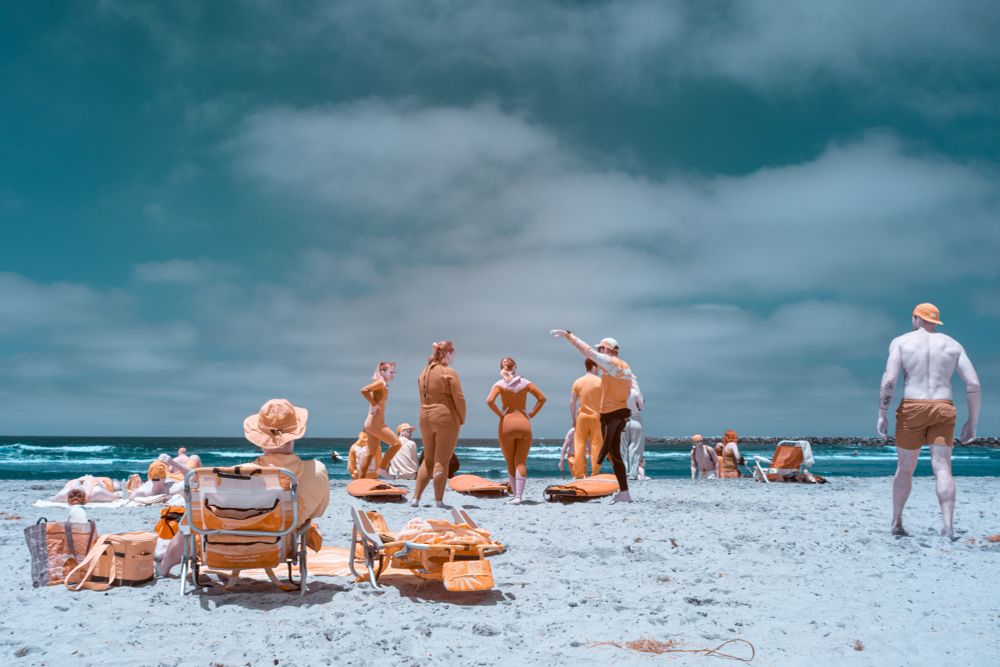 A surf class gathers on the beach, with most participants facing and listening to an instructor who gestures toward the ocean. The group wears warm-toned wetsuits and clothing that appear orange due to infrared capture. A man with pale infrared-exposed skin walks into the frame from the right. Surfboards and beach gear are scattered in the sand. The sky is partly cloudy and the ocean is visible in the background.