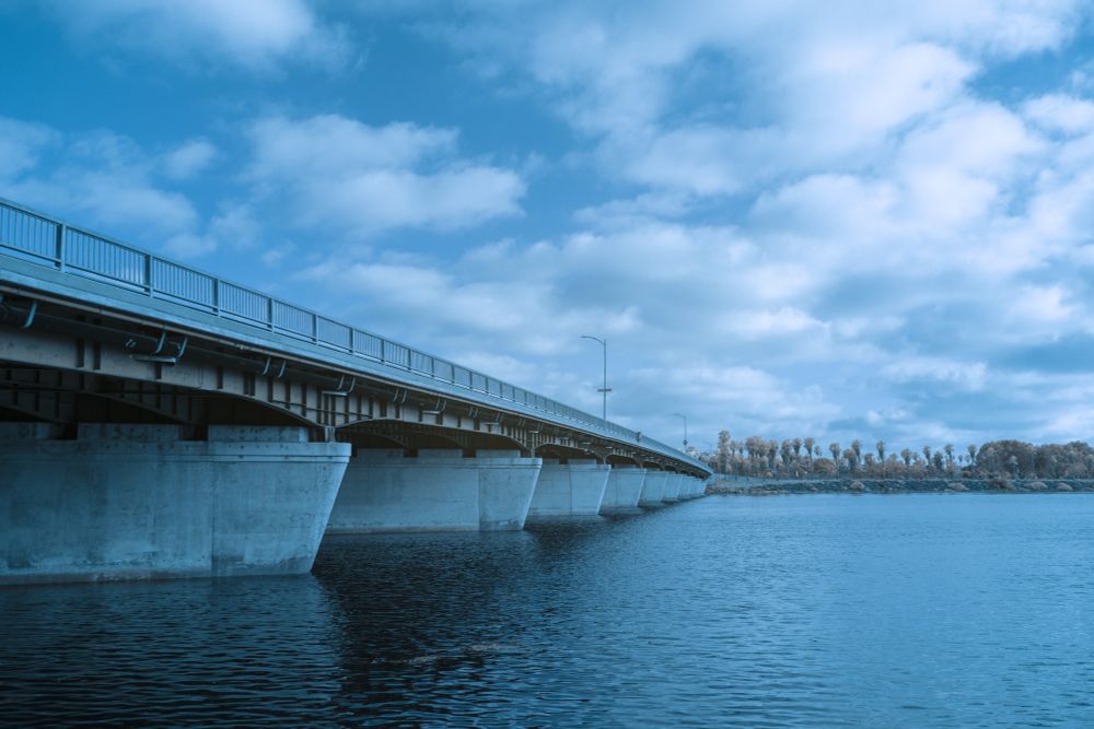 A long concrete bridge extends across a calm body of water under a partly cloudy sky. The image has been processed using infrared color swapping, giving the entire scene a cool cyan-blue cast. Trees on the far shore, including a row of palms, appear in muted tones, contrasting subtly with the strong geometric lines of the bridge. The composition emphasizes depth and vanishing point perspective.