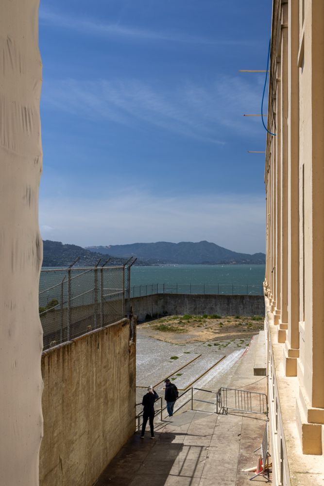 View from Alcatraz Island showing two people walking down a ramp between beige prison buildings. A barbed-wire fence and retaining wall enclose the area, with San Francisco Bay and a distant green hillside visible under a clear blue sky.