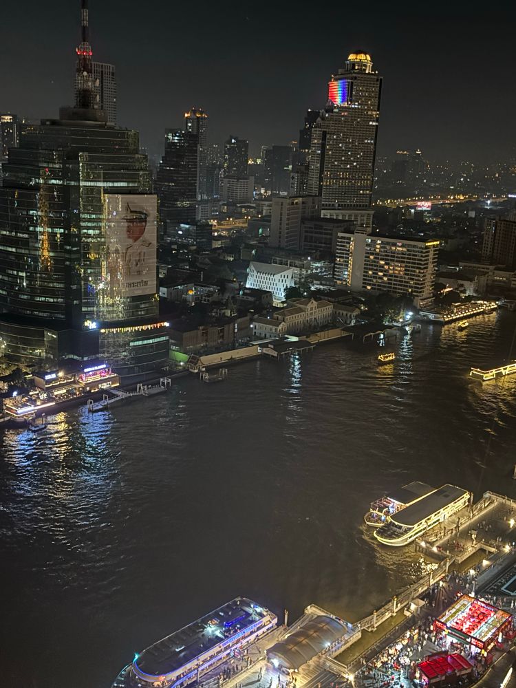 River with boats and skyscrapers in Bangkok. One building is lit up with the colours of the rainbow 