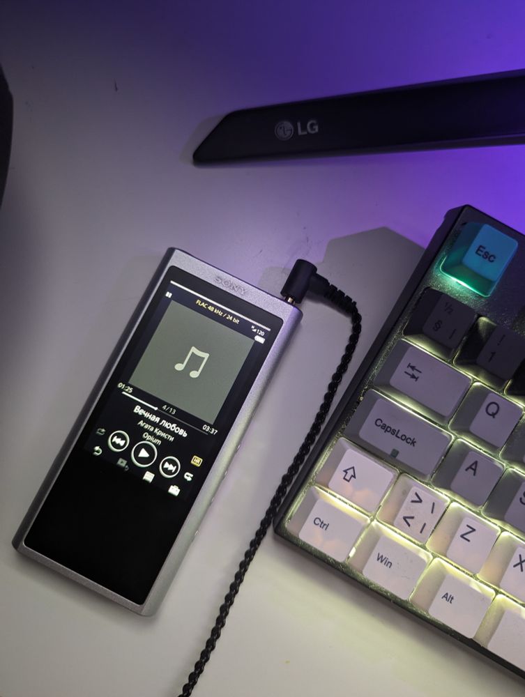 A birdeye view photo of a Walkman NW-ZX300 device on a white table by a keyboard and monitor stand with a headphone cable connected on top. 