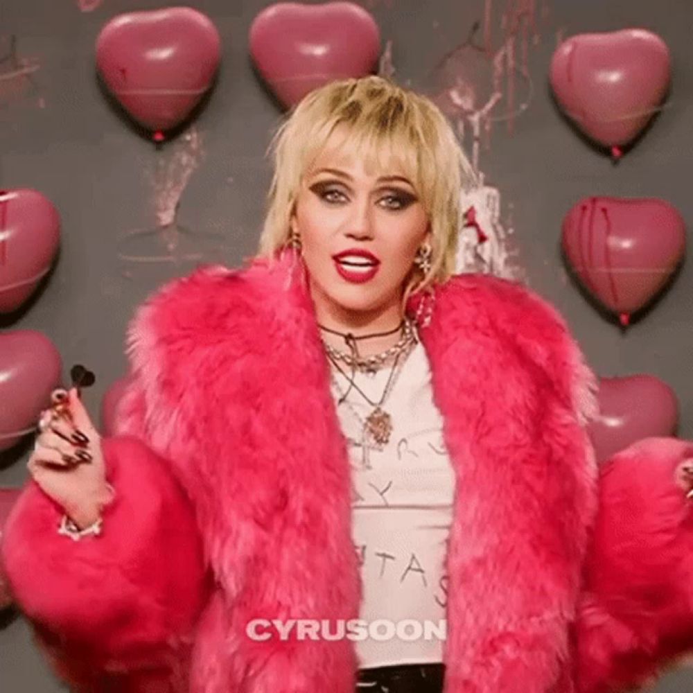 miley cyrus is wearing a pink fur coat and standing in front of a wall of pink balloons .