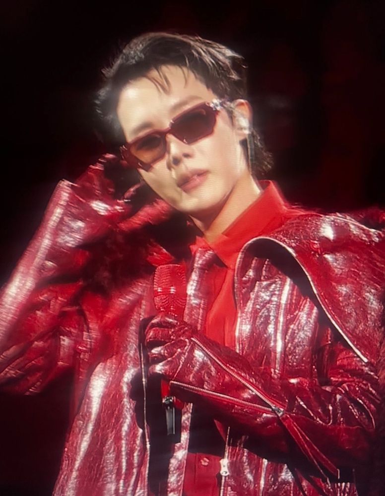 J-hope/Jung Hoseok of BTS on stage in an all red leather or pleather outfit with red shirt and red necktie, gloves, sunglasses, and slicked to the side hair. He's holding a red mike and holding a hand up to his ear. It's slightly blurry, clearly a fan shot