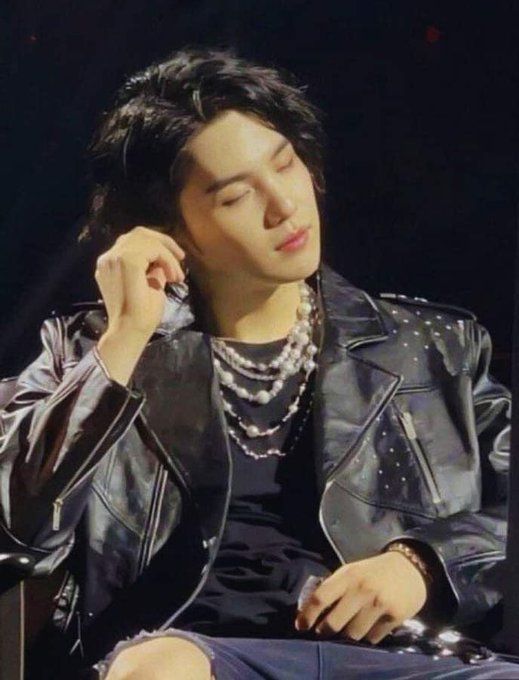 Yoongi/Suga/AgustD of BTS in a black leather jacket and black t-shirt and black ripped jeans sitting down on stage with a mic in one hand and eyes closed, apparently adjusting his in-ear