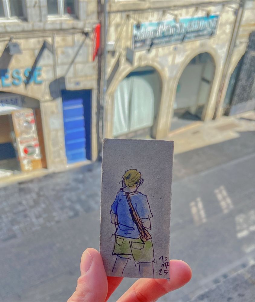 A photo of a street seen from above, with a hand holding a 8x4cm piece of carton featuring a watercolor sketch of a lady walking on the street. 
