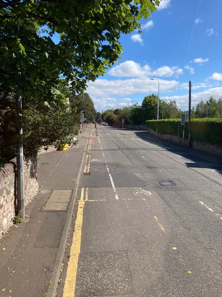 View from top deck front seat of a bus driving down single lane road edge of Edinburgh surrounded by trees.