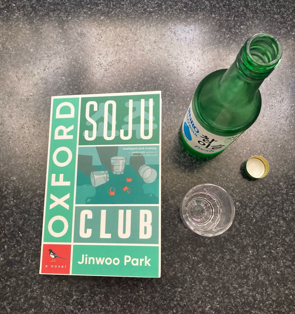 A copy of Jinwoo Park’s novel Oxford Soju Club sits on a black countertop next to a green bottle of soju and a shot glass.