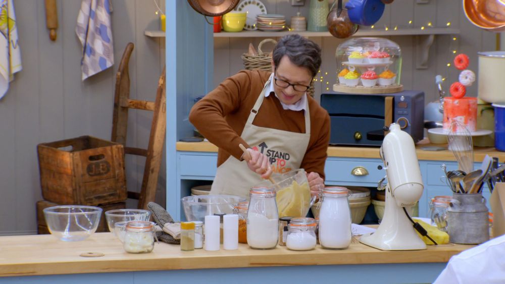 a screenshot of Reece Shearsmith in Great British Bake Off: Stand Up To Cancer. he's wearing a rusty orange sweater, which is on top of a white collared shirt and an apron on top of that, and he's mixing biscuit dough. lots of grey is visible in his floofy hair
