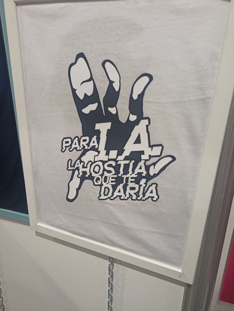Una camiseta blanca colocada sobre un marco de cuadros con un texto de "Para IA la hostia que te daría" con un dibujo de una mano anatómicamente mal (dedos de más, dedos que se doblan donde no tocan, etc).