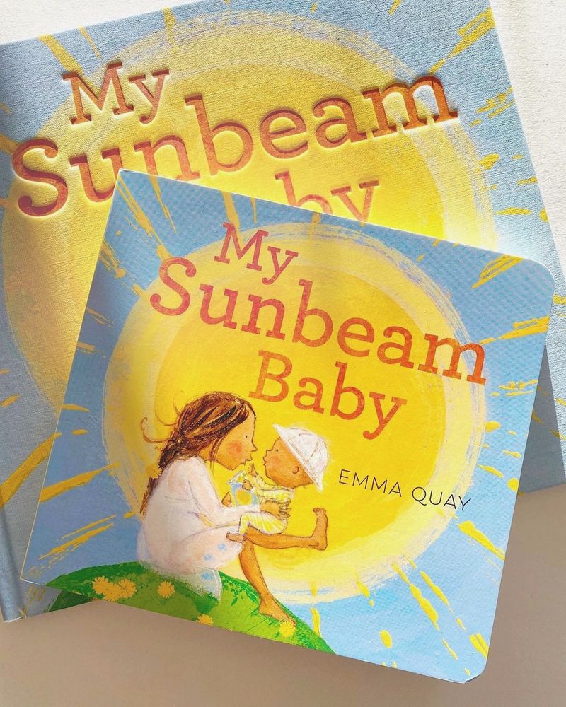 Two copies of the picture book MY SUNBEAM BABY by Emma Quay — the hardback and the board book. Cover of the picture book MY SUBEAM BABY by Emma Quay, in hardback. The cover illustration shows a mother bouncing her baby on her knee, sitting on a hilltop with the sun behind them.