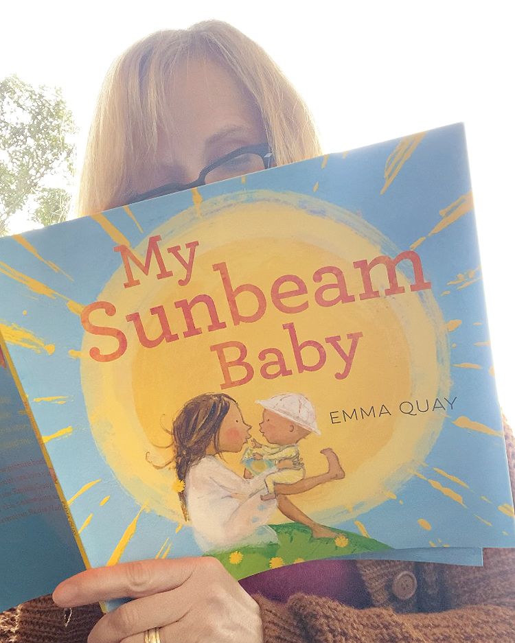 Author-illustrator Emma Quay reading the new paperback edition of her picture book MY SUNBEAM BABY. The cover illustration shows a mother bouncing her baby on her knee, sitting on a hilltop with the sun behind them. 