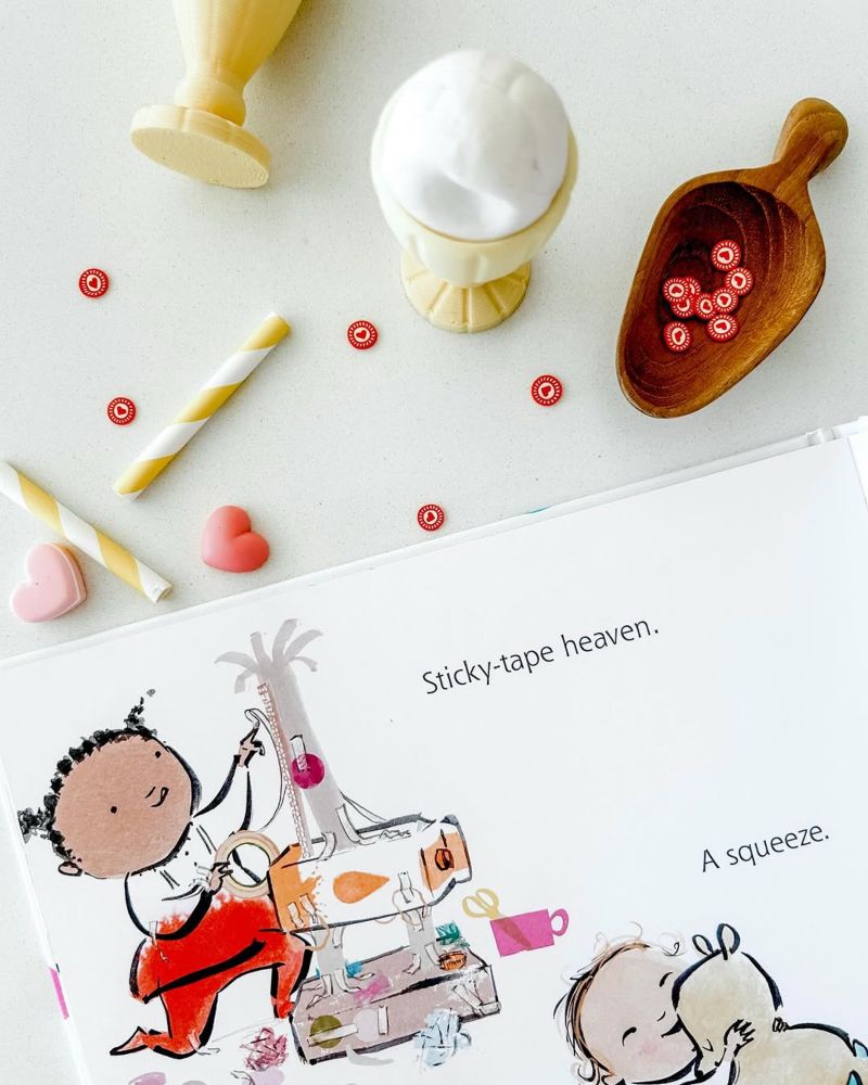The picture book “Happy All Over”, open to a colourful illustration by Emma Quay and with ice-cream sundae toys and confetti scattered around.