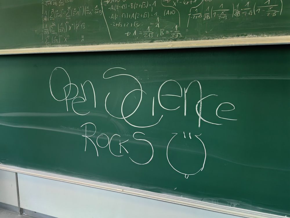 A chalkboard with the words "Open Science Rocks" written in white chalk with a smiley face drawn next to it. Complex mathematical equations left from lectures prior to the event are slightly visible on the chalkboard above it.