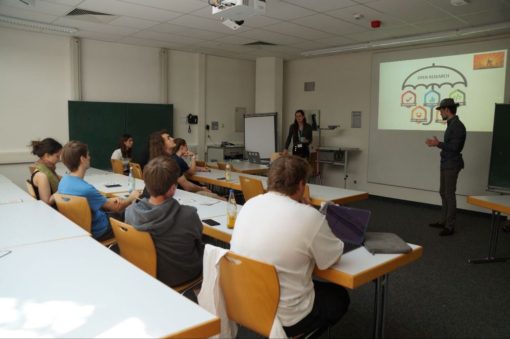 Two presenters lead a discussion in a classroom, with a projected slide showing icons related to Open Research, while students seated at desks listen.