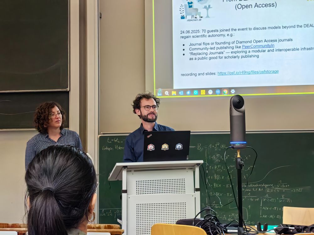 Man (Felix Schönbrodt) and woman (Malika Ihle), both with curly hair, glasses and blue shirts, stand beside a laptop and a conference camera at the front of a lecture room giving a presentation. A slide on "Open Access" is projected behind them. The back of an audience member’s head is visible.