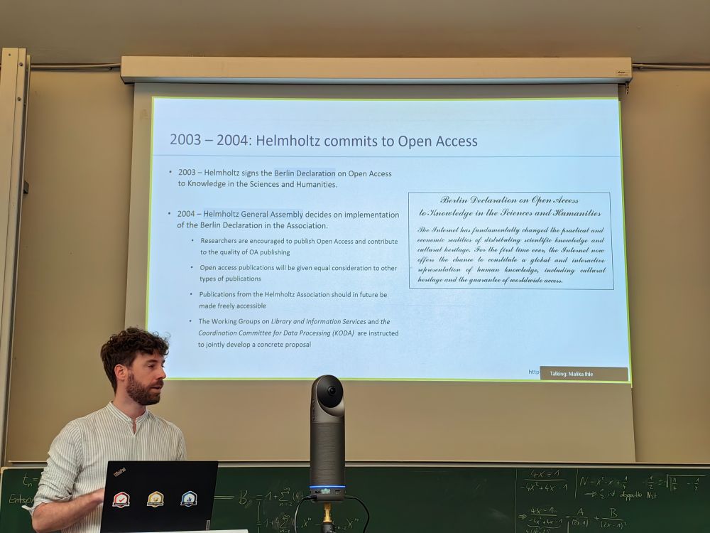Presenter (Dr. Mathijs Vleugel), a young, white man with a beard wearing a beige shirt, stands beside a laptop and a conference camera in front of a projection screen. The slide behind gives information related to Helmholtz’ commitment to Open Access in 2003–2004.