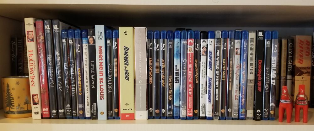 Shelf with copies of Holiday Inn, a Bing & Frank holiday special, I'll Be Seeing You, Since You Went Away, The Lion in Winter, It Started With Eve, Paddington 2, the Gerwig and Armstrong versions of Little Women, Meet Me in St. Louis, Road to Utopia, Battleground, Remember the Night, Fanny and Alexander, A Tale of Two Cities, The Thin Man, After the Thin Man, When Harry Met Sally, The Thing, Cranford, White Christmas, On Dangerous Ground, Planes Trains & Automobiles, Slap Shot, The World of Henry Orient, Auntie Mame, The Shop Around the Corner, It's a Wonderful Life, The Muppet Christmas Carol, The Night of the Hunter, Donovan's Reef, The Apartment, Stalag 17.