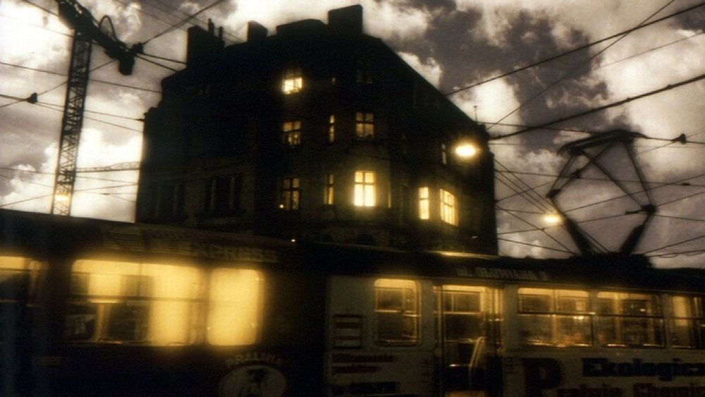 C'est une image sépia tirée du film : un train passe devant un grand bâtiment au crépuscule. Le ciel est nuageux et de la lumière jaune émane des fenêtres du train et du bâtiment.