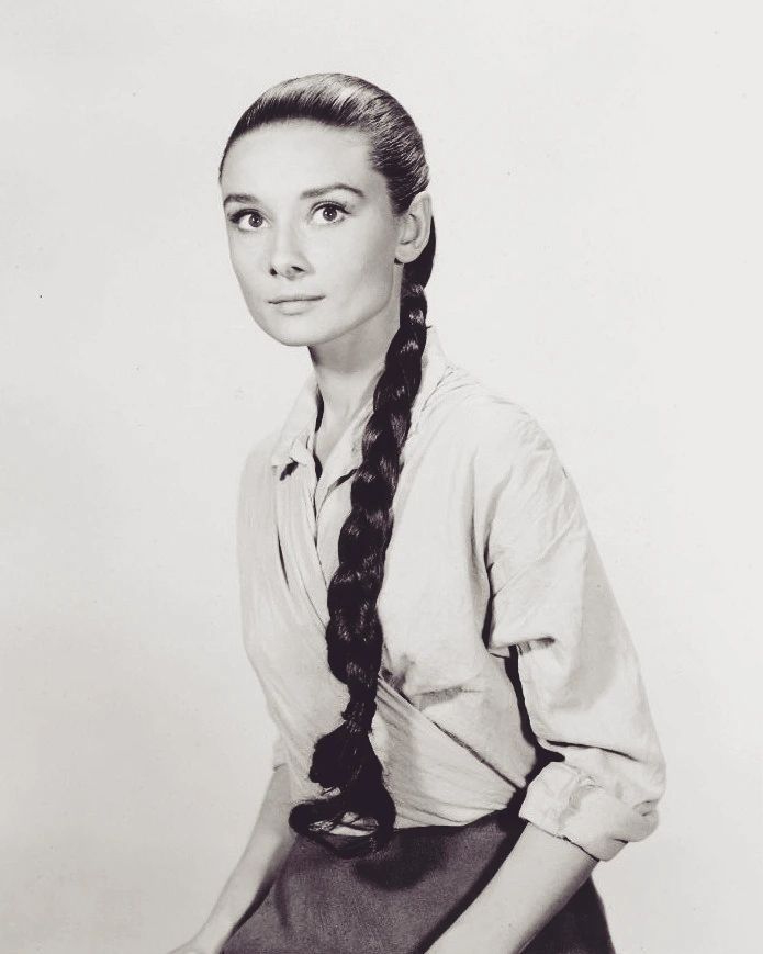 Une photo d'Audrey Hepburn en 1959 sur le tournage du Vent de la Plaine de John Huston : c'est une femme blanche d'une trentaine d'année avec une chemise blanche à manches retroussées et de longs cheveux bruns rassemblés en une natte sur son torse.