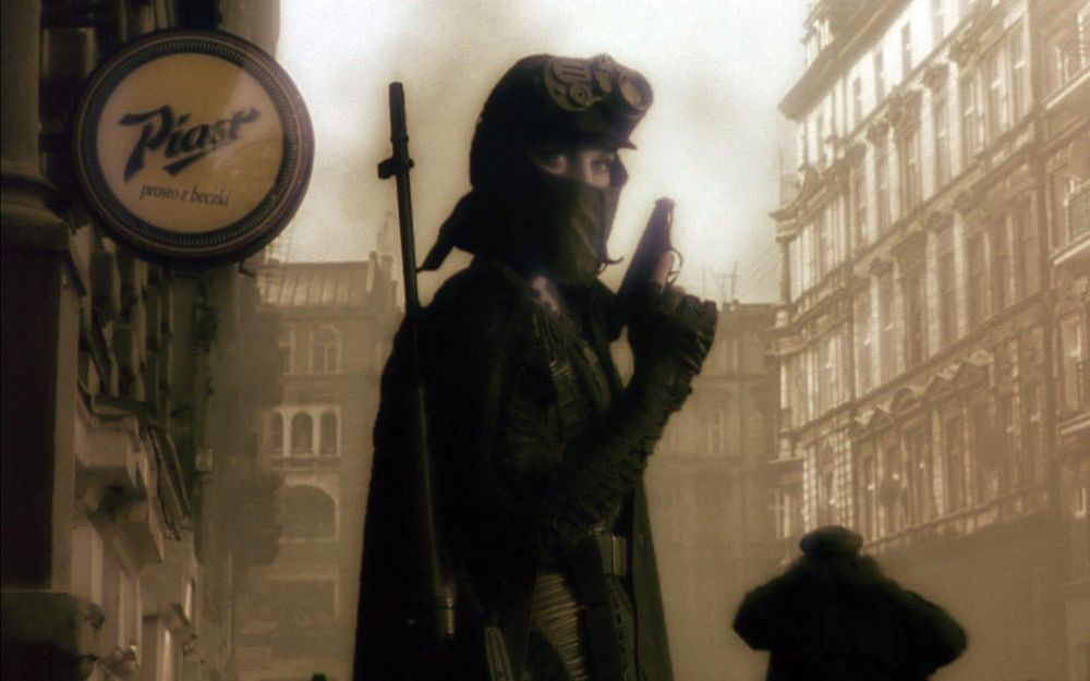 C'est une image sépia tirée du film : une femme masquée, avec des lunettes de vision nocturne sur le front, un pistolet en main et un fusil en bandoulière, se tient dans au pied de bâtiments à l'architecture européenne. Une enseigne ronde qui indique "Plast" est accrochée derrière elle.