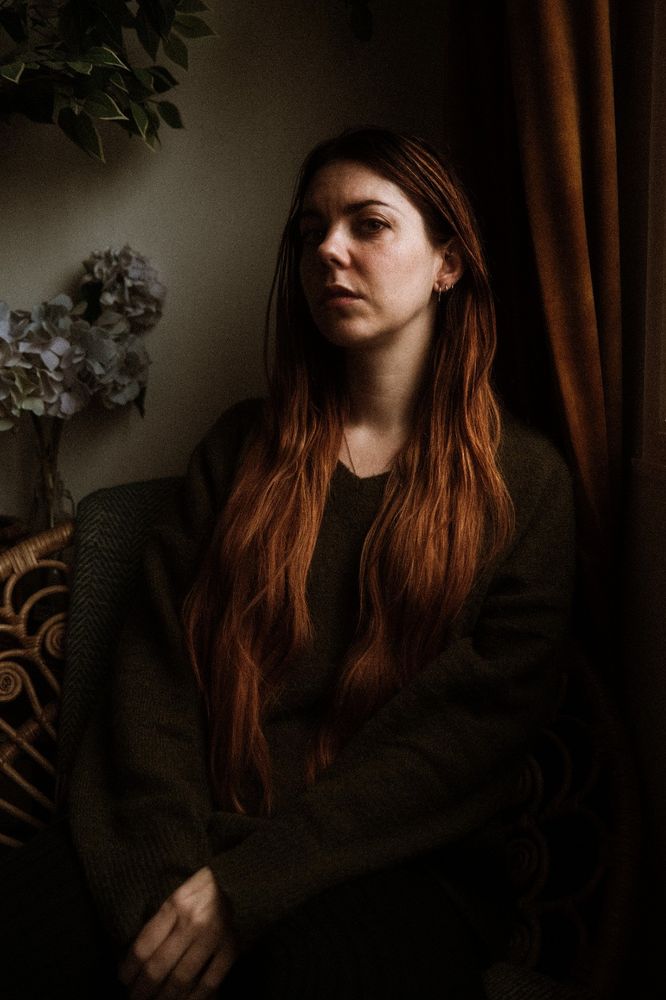 Portrait où je suis assise sur une chaise en rotin, il y a un rideau ocre à droite et des fleurs bleu-violet à gauche. L'ambiance colorée a de l'ocre un peu chaud et du vert foncé comme la forêt 