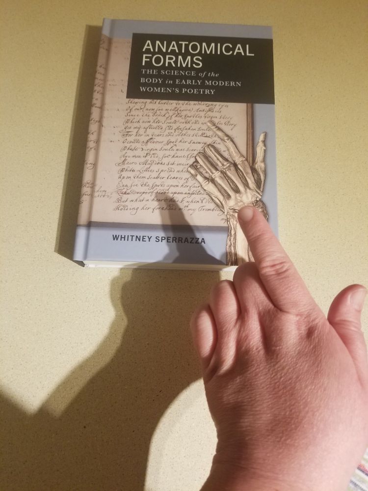 A picture of a book entitled Anatomical Forms: The science of the body in early modern women's poetry, by Whitney Sperrazza. The cover has a skeletal hand pointing at a page of manuscript poetry, and in the foreground a human hand points at the skeletal hand, mimicking the cover.