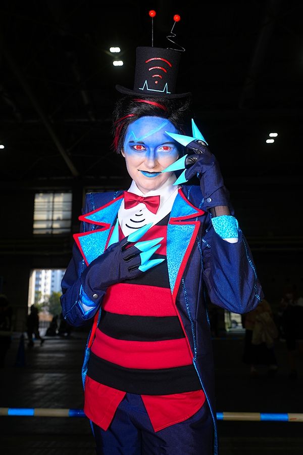 Rachel is cosplaying a gijinka version of Vox from Hazbin Hotel. She's standing with her right hand to her chest and left hand beside her face in electric blue clawed gloves. The lighting is stark, and the background is mostly dark.

She wears a short black wig with streaks of red. The top half of her makeup is bright blue, like Vox's screen, with screen glare painted across her forehead. From the bridge of her nose to her chin is a natural white skin tone, save for a striking blue lip for her satisfied smirk. 