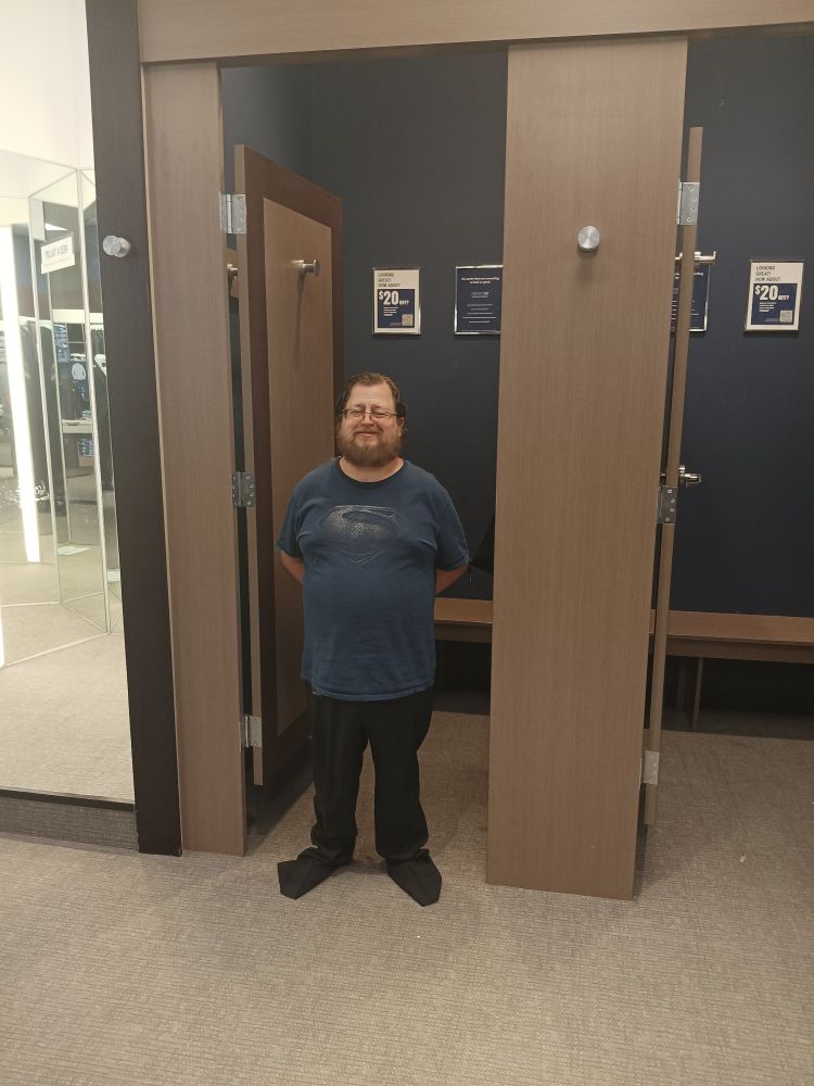 Man in superman shirt with trousers on that are too big standing proudly in the doorway of a changing room
