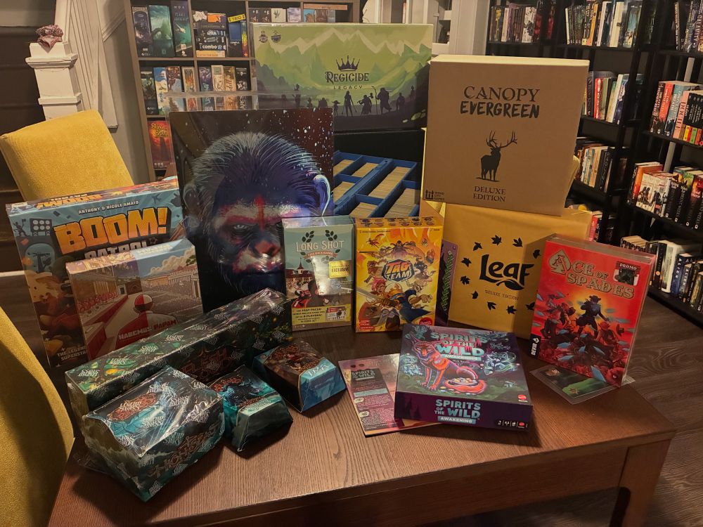 A dinner table with a mixture of board games, card games, and art pieces. Boom! Patrol, Canopy Evergreen, Leaf, Ace of Spades, Spirits of the Wild: Awakening, Tag Team, Long Shot, Regicide Legacy, and several products from Flesh and Blood TCG as well as a painting of Caeser from the new Planet of the Apes films.