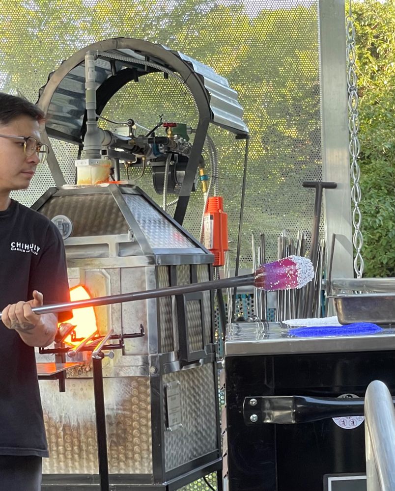 A man in front of a glass furnace holding a metal pipe with a gob of molten glass on the end, rolling the glass in shards of coloured glass
