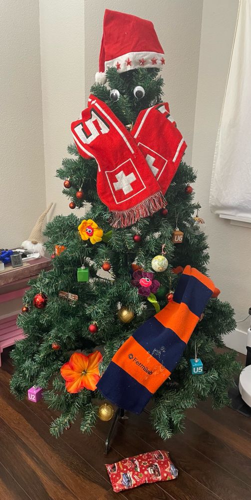 The same tree as in the replied-to post, but now with a red and white Swiss scarf around the ‘neck’ of the tree (just under the googly eyes) and a blue and orange Fermilab scarf reaching partway around the bottom. 