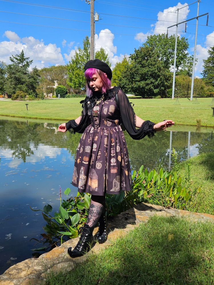 Blix in front of a manicured lawn and pond. Their hair is magenta today. They're wearing a black dress with a print of assorted spooky houses and especially stupid shoes with curling witch toes.