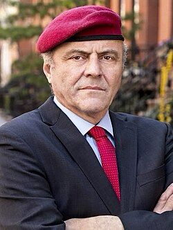Picture of Curtis Sliwa from his Wikipedia page. He is wearing a dark suit with a red tie and a red beret flopped over to the side