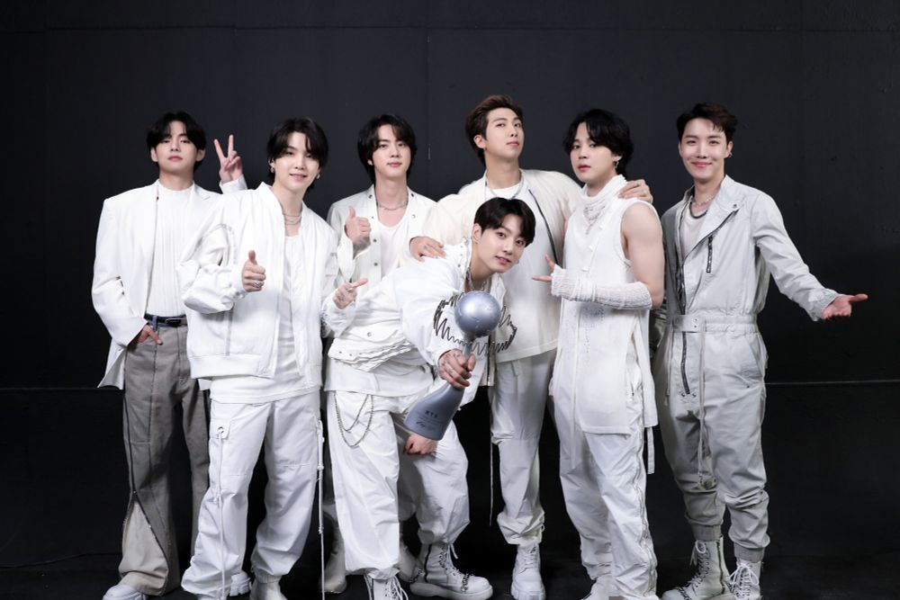 bts ot7 backstage at a 2021 ptd live on stage concert. they're wearing their white and gray opening outfits, cyberpunk assassins meets space fashion models. jungkook is leaning over in front, holding out what looks like a giant squiggly upside down exclamation point with a squiggly halo to us. this is actually bts's IFPI global recording artist of the year award. he looks quite proud.