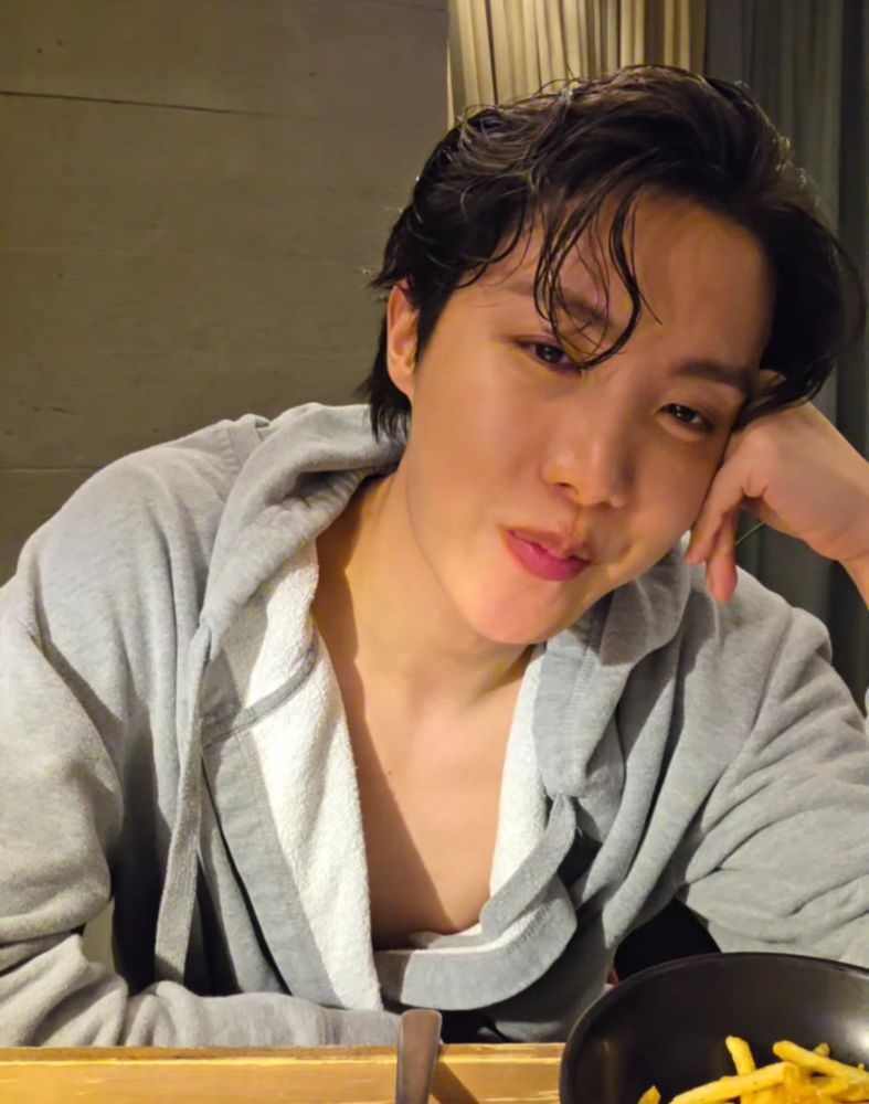 j-hope on weverse live in a loose bathrobe, leaning his head on his hand, but this time looking into the camera and smirking a little.