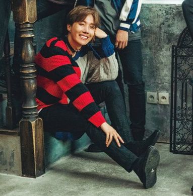 hobi, in the same outfit again, sitting on the bottom step of a staircase, head resting on his hand and smiling brightly. he's gorgeous.