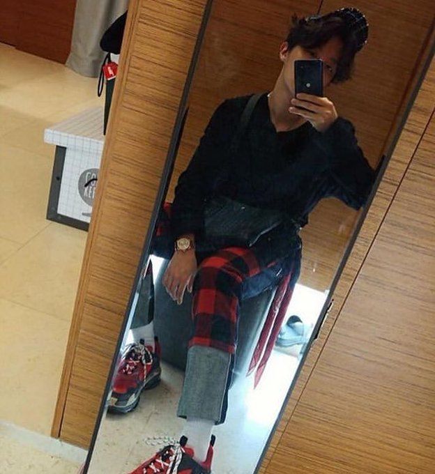 j-hope in 2018, wearing jeans with red flannel patches, red balenciaga sneakers, and a baseball cap. sitting on a stool reflected in a narrow full length mirror. his phone covers most of his face. his watch is prominently displayed.