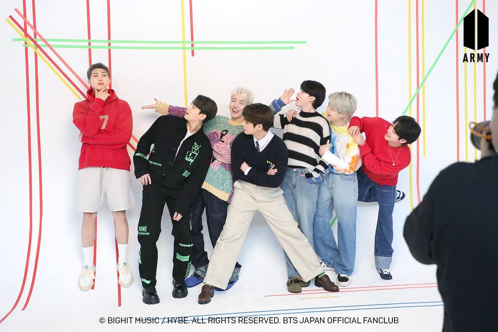 bts ot7 from a 2021 japan official fanclub shoot. they're posing against a studio backdrop of white with colored lines. namjoon at one end has one eyebrow raised and fingers thoughtfully on his chin, standing a bit higher than the rest. everyone else is leaned over to look at him properly, admiring him in all his namjoon-ness.
