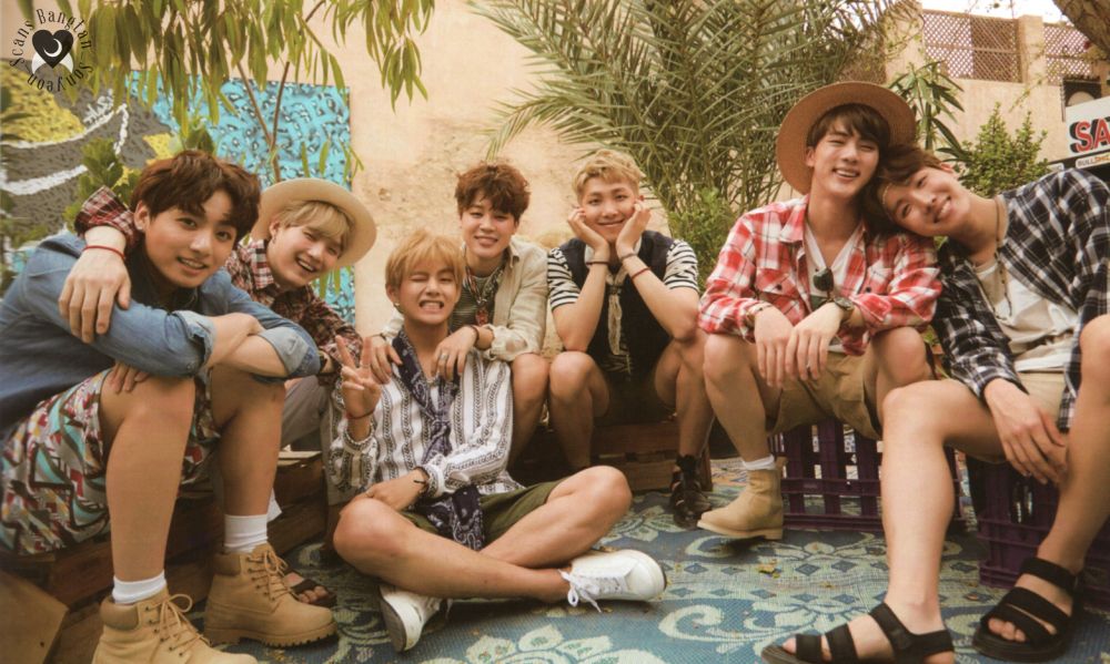 bts ot7 for the 2016 summer package in dubai. they're sitting on a very low ledge almost on the ground, in lots of shorts and light casual clothing, cuddled happily together. hobi is resting his head happily on jin's shoulder, taehyung is leaning back against jimin's knees, yoongi's arm is around jungkook, and namjoon is cheesing with his cheeks in his hands.