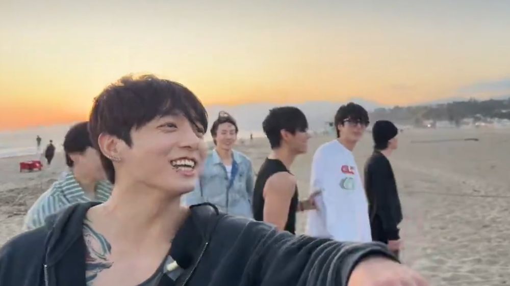 jungkook on the beach on weverse live today, his hoodie fallen open to reveal some of the tattoo on his chest. a mic is taped to the opposite inside edge of the hoodie.