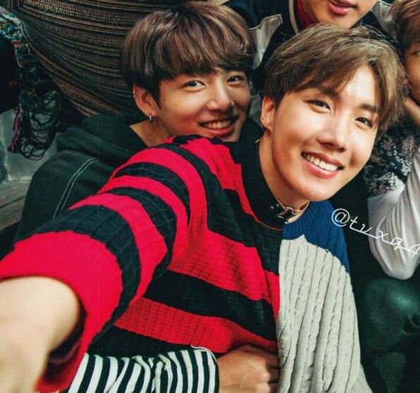 hoseok, in the same outfit, smiling with one arm out as if taking a selfie. he looks radiant. behind him, jungkook is holding him close with one arm around his middle. 