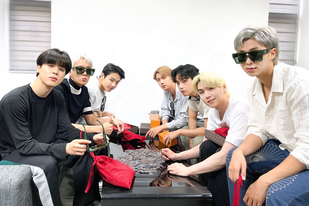 bts ot7 in 2021. they're seated around a table filled with puzzle pieces that will form their invitation to a louis vuitton fashion show. they're dressed in regular casual clothes but both hobi and namjoon are wearing ridiculous giant square louis vuitton sunglasses and somehow pulling them off. hobi's hair is short and white-blonde with tiny bangs, and jimin has a sleek black bowl cut.