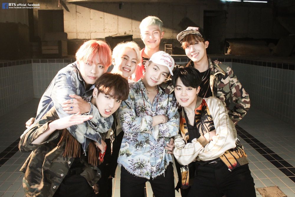 bts ot7 in 2016 on the set of the fire mv. they're standing in an empty indoor pool, dressed in camo and tropical prints, posing for the camera. yoongi and jimin have their arms crossed like they're tough, jungkook is making a snarly face and karate chopping at the camera while taehyung clings to his back, and the rest of them are cheesing.