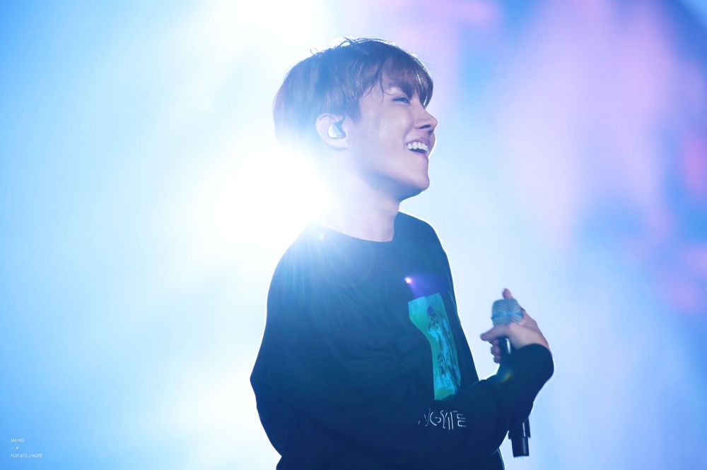 j-hope on stage at the pyeongchang winter olympics concert in 2016. he's in profile, smiling cheekily, with stage light flooding in behind the back of his neck. he looks like he's having the time of his life.